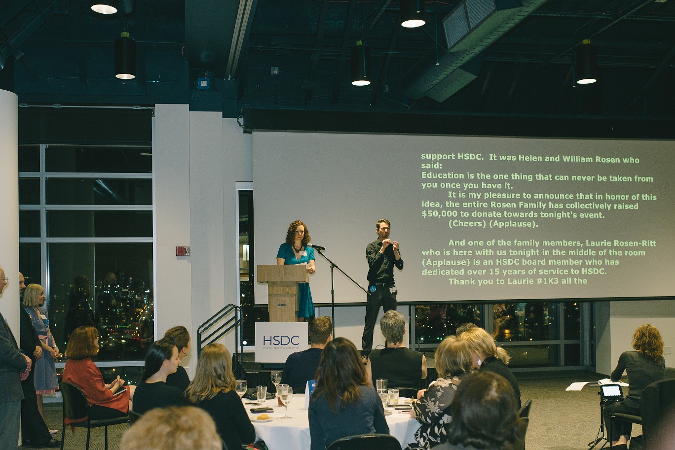 A speaker and interpreter at HSDC's 2017 Cocktails & Connections event, with live captioning behind them.