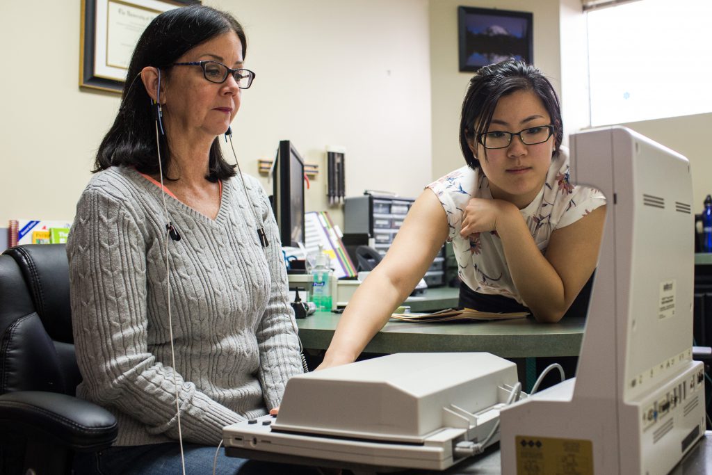 A photo of an audiology appointment. A client sits on the left with a device in front of them. On the right, an audiologist reaches over their desk to adjust the device.