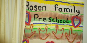 A sign reading "Rosen Family Preschool". Underneath the text is the outline of hands making the ASL sign for "I love you".