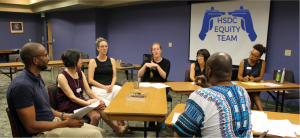 The HSDC Equity Team meeting in August 2019. Seven people sit around a U-shape of tables in a conference room. The HSDC Equity Team logo is on a projector screen.