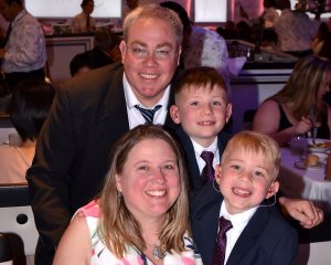 The Gramer family posing for a picture in a restaurant. Father Brendan, mother Laura, and brothers Brett and Luke. They are wearing formal attire.