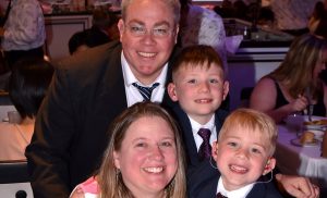 The Gramer family posing for a picture in a restaurant. Father Brendan, mother Laura, and brothers Brett and Luke. They are wearing formal attire.