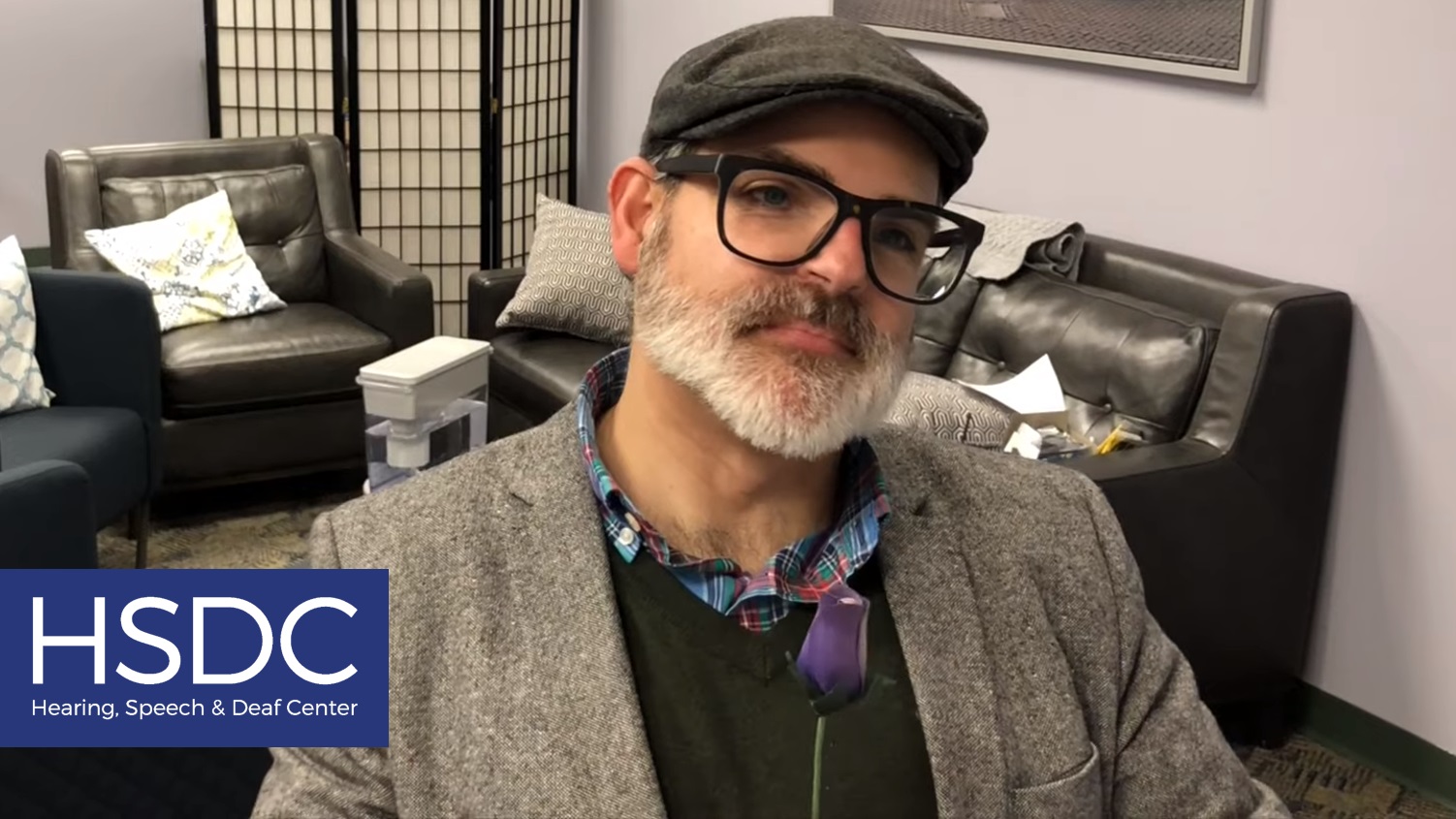 A screenshot of a video. A man with a gray beard, black glasses, grey hat, and grey coat is holding a blue flower and looking offscreen to the right. The HSDC logo is in the bottom left corner.