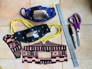 A photo of three face masks on a tile floor. The two finished masks on top have clear plastic rectangles around the mouth area. The unfinished mask on the bottom has a hole where the plastic will go. On the right are a metal ruler, Sharpie, and scissors.