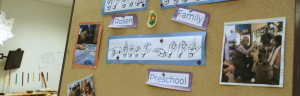 A photo of a corkboard in Rosen Family Preschool. There is paper with the school name, as well as a paper with ASL spelling PRESCHOOL. The name is surrounded by a few photos of the teachers and students in class.