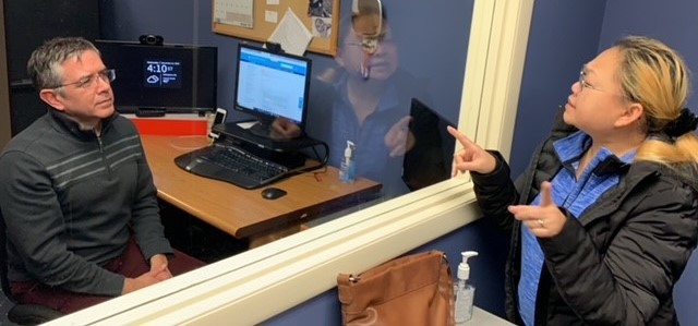 A photo of HSDC client Thuy signing with HSDC Client Advocate Dino. They are talking from opposite sides of a glass window indoors. On the right, Thuy is standing and signing. On the left, Dino is sitting and watching. Thuy has tan skin with blonde hair. Dino has tan skin, and short salt and pepper hair.