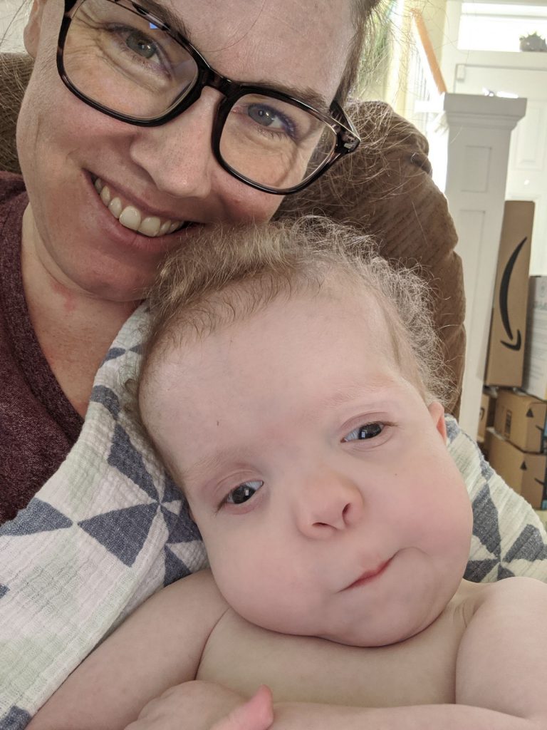 Levi and Mom sitting in a chair, smiling at the camera. Mom is holding Levi in a blanket. Levi and Mom are both light-skinned. Levi has light brown hair. Mom has brown hair and glasses.