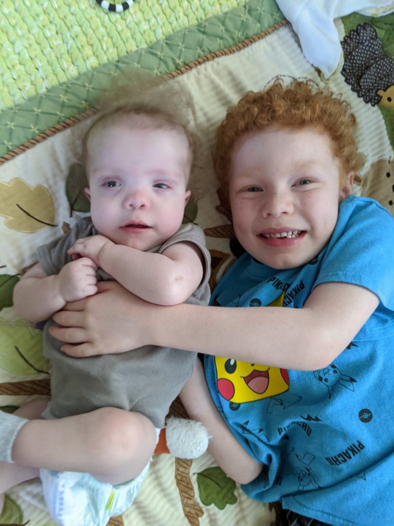 Levi and an older sibling lying on a blanket on the floor looking up at the camera. The sibling is lightly hugging Levi. Both are light-skinned. Levi has light brown hair, and wears a green onesie. The sibling has curly red hair, and wears a light blue t-shirt.