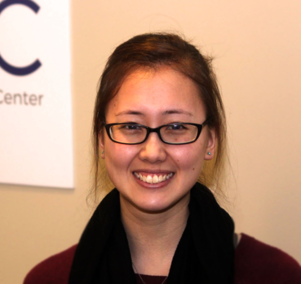 HSDC staff member Keito Omokawa smiles for the camera. Keito is light-skinned female, with brown hair, pull back in braids. She is wearing glasses and black scarf over maroon sweater.