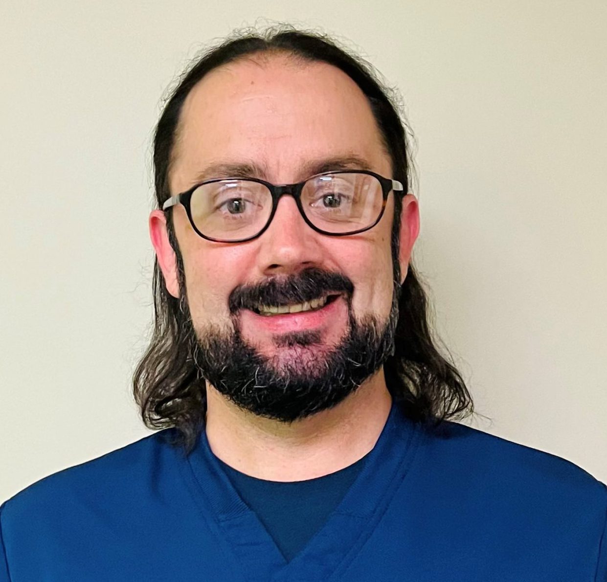 HSDC staff member Chris Stefanile smiles for the camera. Chris is male, with dark hair, pull back in ponytail. He is wearing glasses and blue scrub.